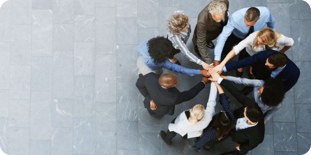 A diverse group of professionals standing in a circle with their hands stacked together in the center, symbolizing teamwork and unity.