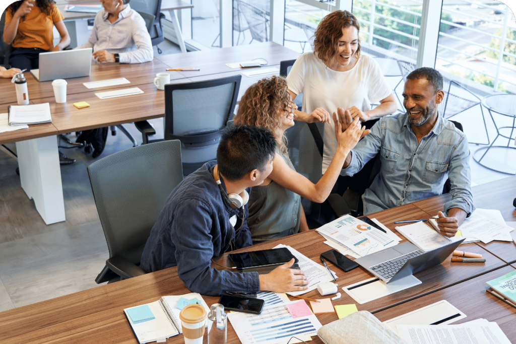 Team celebrating a win around a conference table covered with laptops, charts, and notebooks.